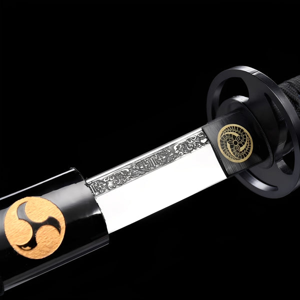 Mitsudomoe Katana close-up showing polished 1060 blade with elaborate fuller engravings, black iron-alloy openwork tsuba with triple tomoe cutout, gold mitsudomoe habaki medallion, and black cotton ito with blue menuki