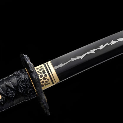 Kurojin Raikiba Katana close-up showing black 1060 blade with white angular lightning engravings, black iron-alloy dragon tsuba, brass honeycomb habaki, and black cotton ito with gold menuki over synthetic rayskin