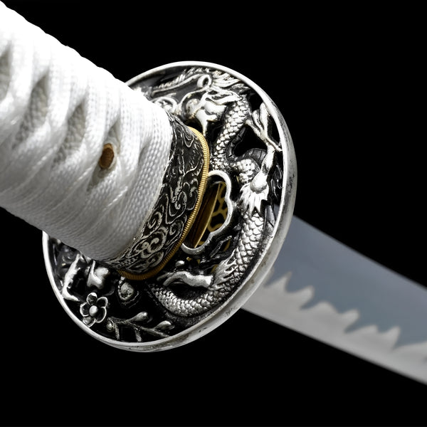 White katana sword close-up showing navy-blue blade with visible notare hamon, brass openwork habaki, iron floral tsuba with silver rim, and white twisted cotton ito